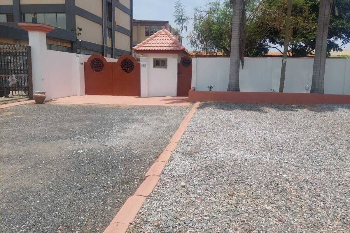 Uma área de estacionamento cercada, com piso de cascalho, um pequeno edifício com telhado de telhas vermelhas e uma parede branca com rodapé vermelho.