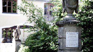 A stone urn with a plaque, surrounded by greenery and a building with arched windows in the background. - Schloss zu Hopferau (Hopferau)