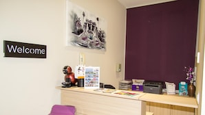 A reception desk with a 'Welcome' sign, a framed artwork, a bulletin board, and a purple chair. - Hostal Barcelona Travel (Barcelona)