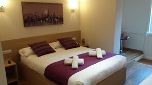 A neatly made bed with a purple throw blanket and two pillows, a nightstand with a lamp, and a cityscape painting on the wall. - Hostal Barcelona Travel (Barcelona)