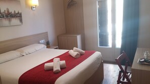 A hotel room with a bed, a desk, a chair, a window with blinds, and a wall-mounted light. - Hostal Barcelona Travel (Barcelona)