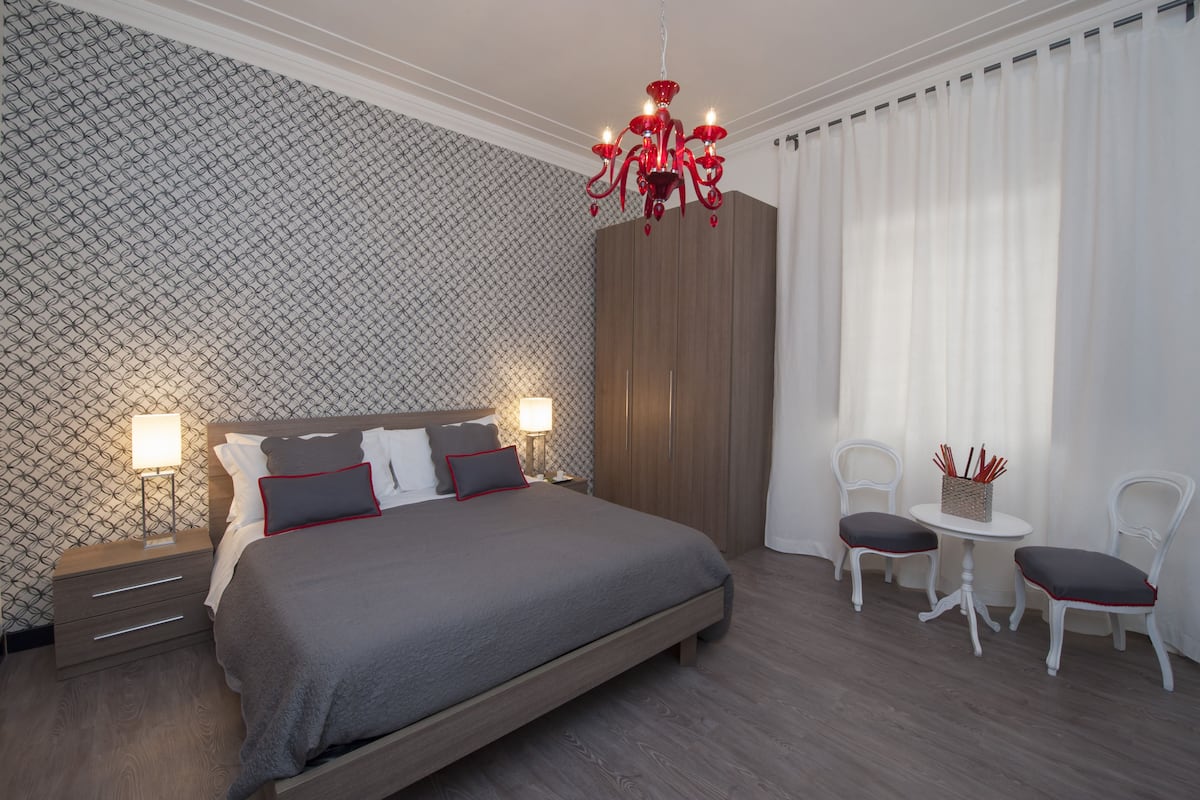 A modern bedroom with a large bed, bedside tables with lamps, a wardrobe, and a seating area with two chairs and a small table.