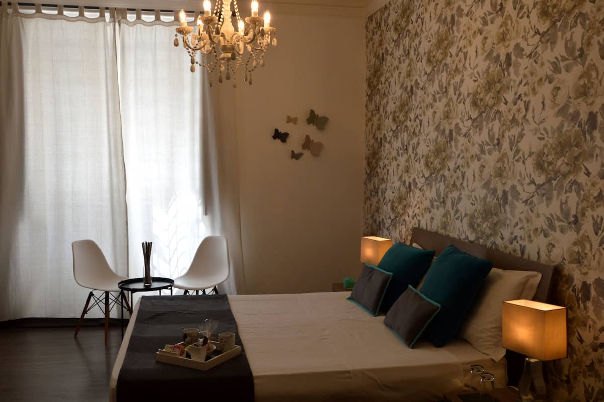A hotel room with a bed, two chairs, a table with a tray of items, a chandelier, and a floral wallpaper.