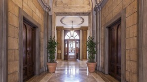 Un amplio pasillo con suelo de mármol, flanqueado por grandes puertas de madera y columnas, que conduce a una entrada imponente con candelabro.