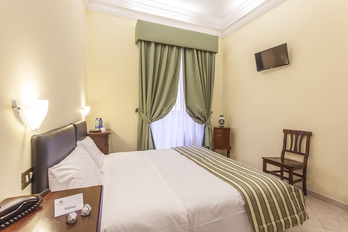 A hotel room with a bed, a bedside table with a telephone, a small table with a welcome card and chocolates, a chair, a nightstand with a lamp, and a TV mounted on the wall.
