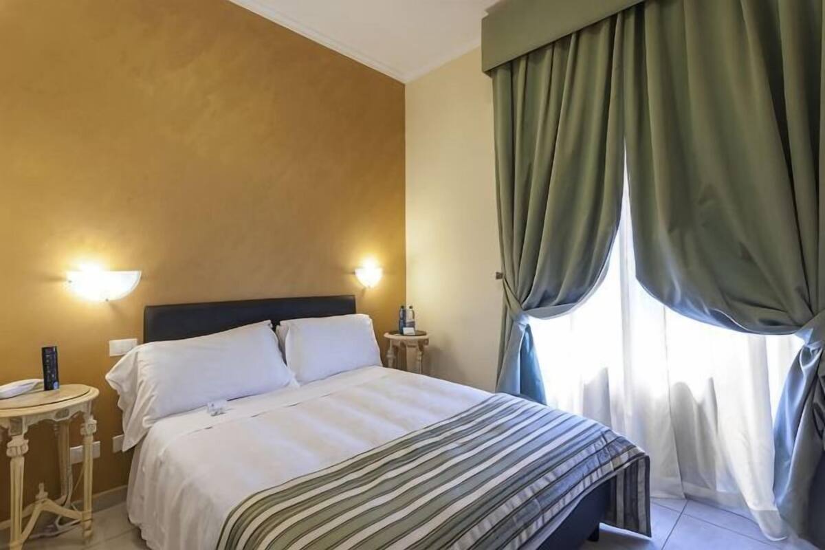 A hotel room with a bed, bedside lamps, a nightstand, and a window with curtains.
