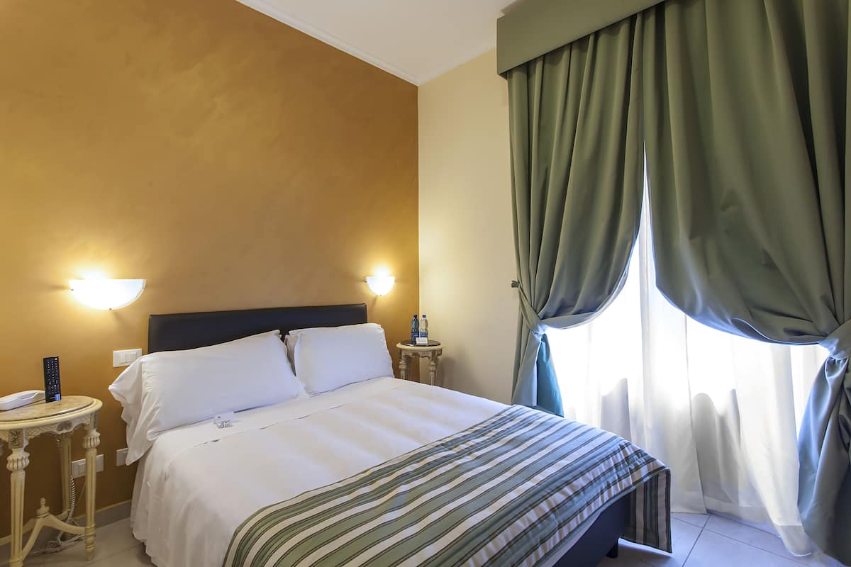 A hotel room with a bed, two bedside lamps, a round table with a phone, and a window with curtains.