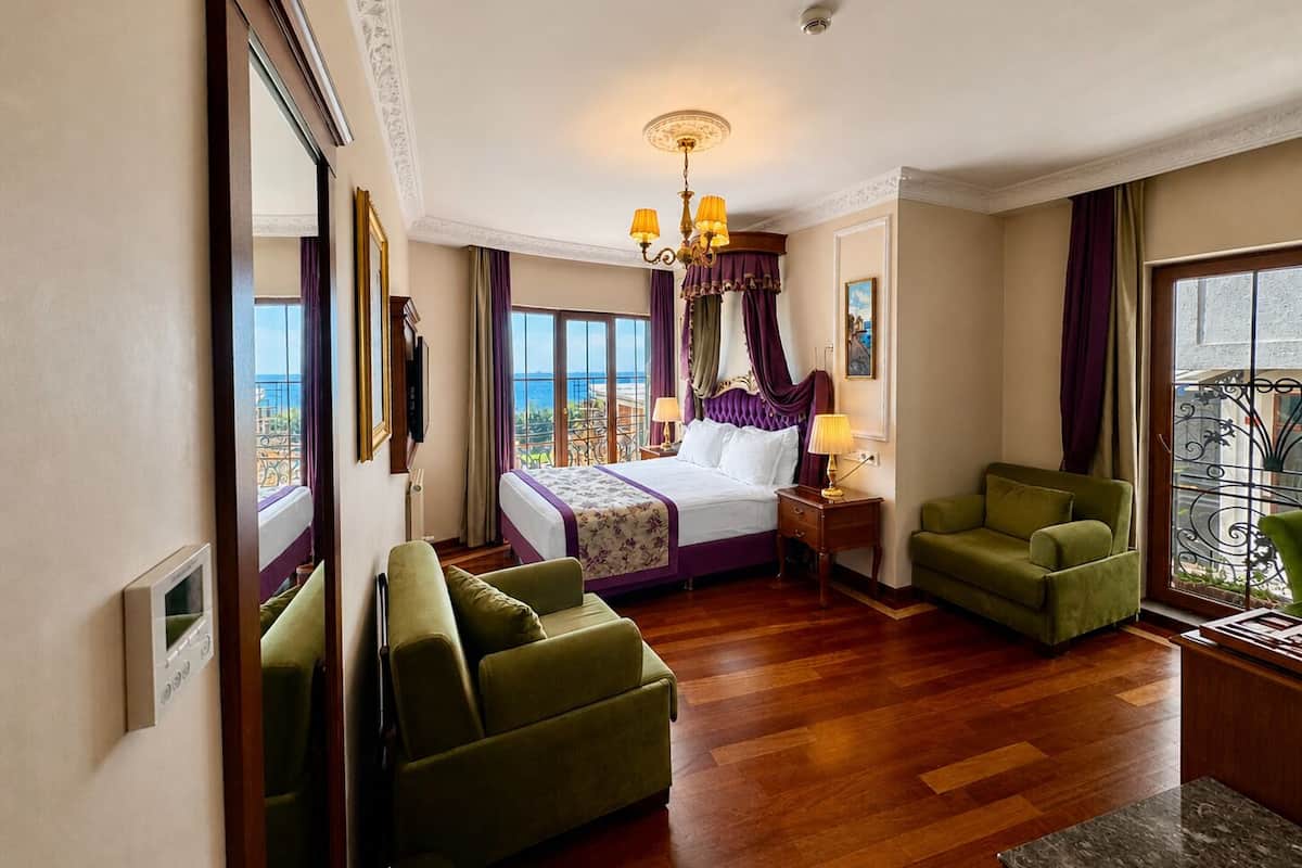 A hotel room with a bed, two green armchairs, a wooden floor, a balcony with a view, and a chandelier.