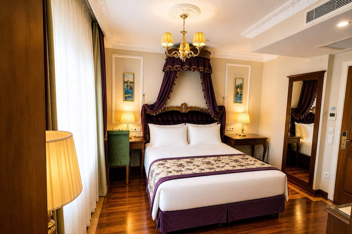 A hotel room with a large bed, a desk, a chair, a chandelier, and a mirror.