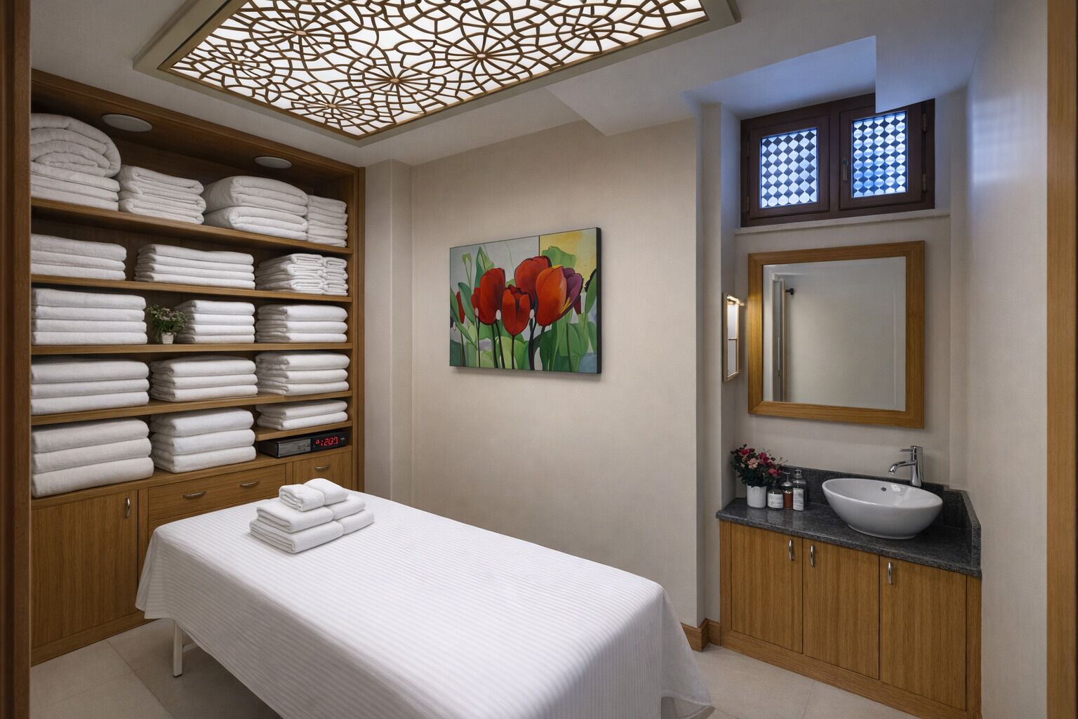 A hotel room with a bed, a large wooden shelf with folded towels, a painting of red flowers, a bathroom with a sink and mirror, and a decorative ceiling light.