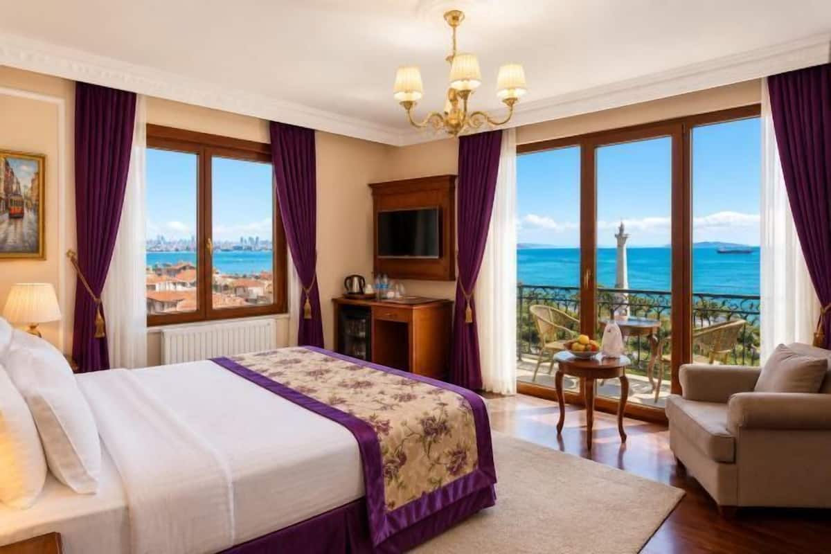 A hotel room with a bed, a TV, a desk, a chair, and a balcony with a view of the sea.