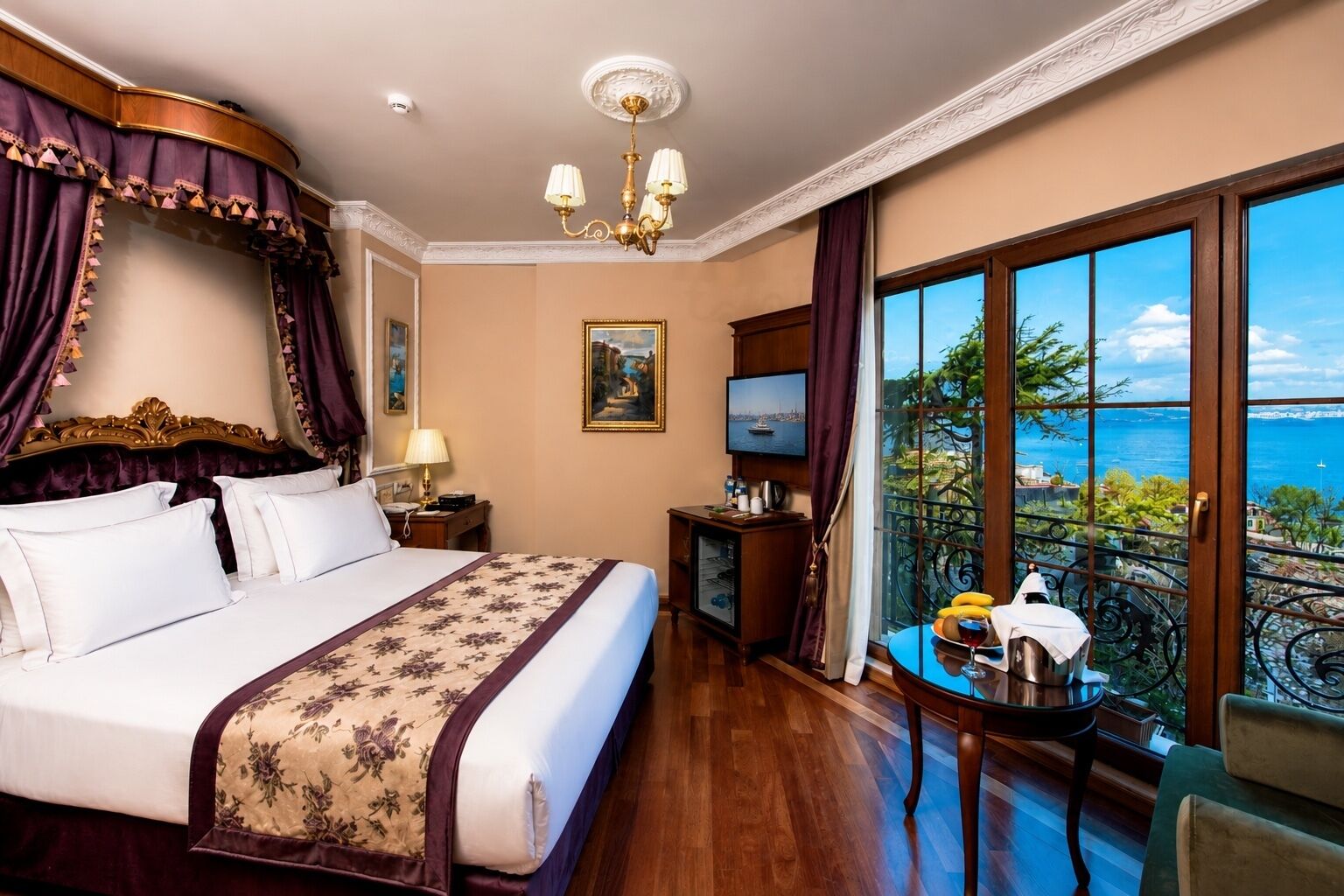 A hotel room with a large bed, a desk with a lamp, a television, and a balcony with a view of the sea.