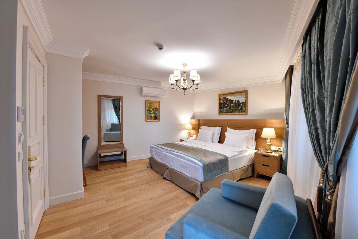 A hotel room with a bed, bedside tables, a desk, a chair, a chandelier, and a painting on the wall.