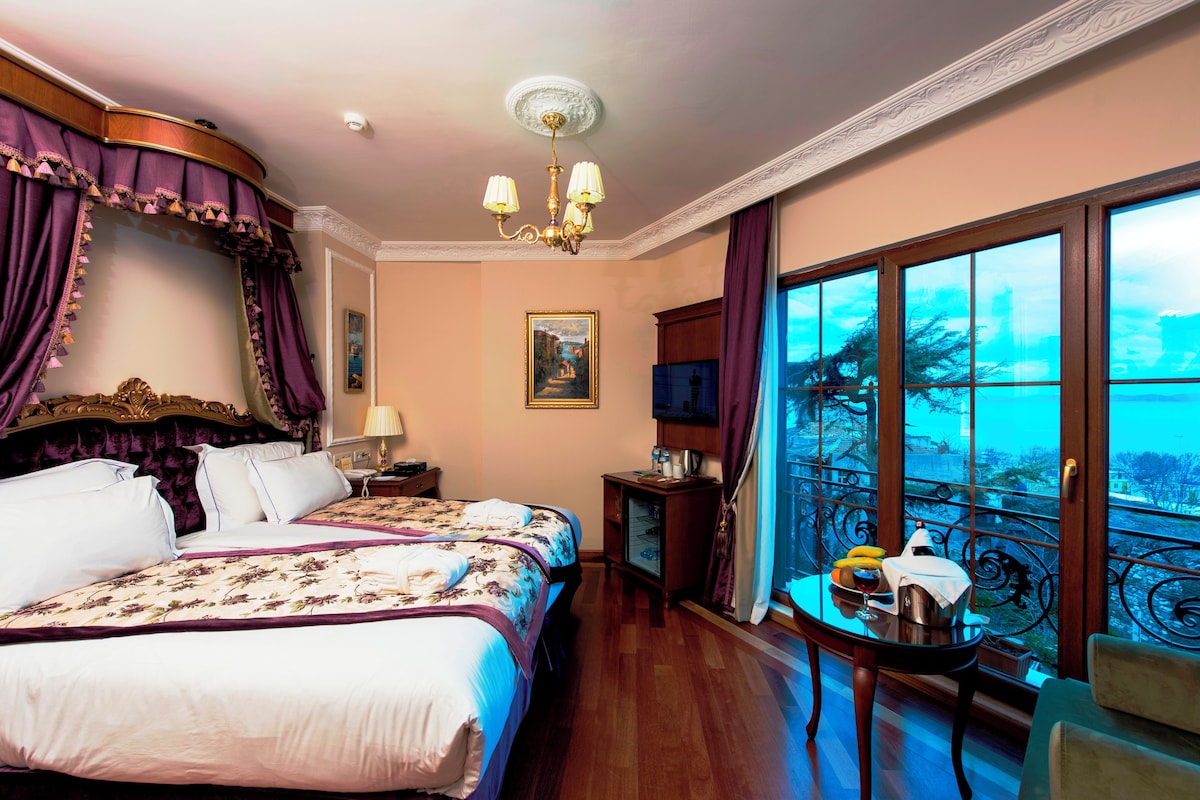 A hotel room with a large bed, a table with a chair, a television, and a balcony with a view.