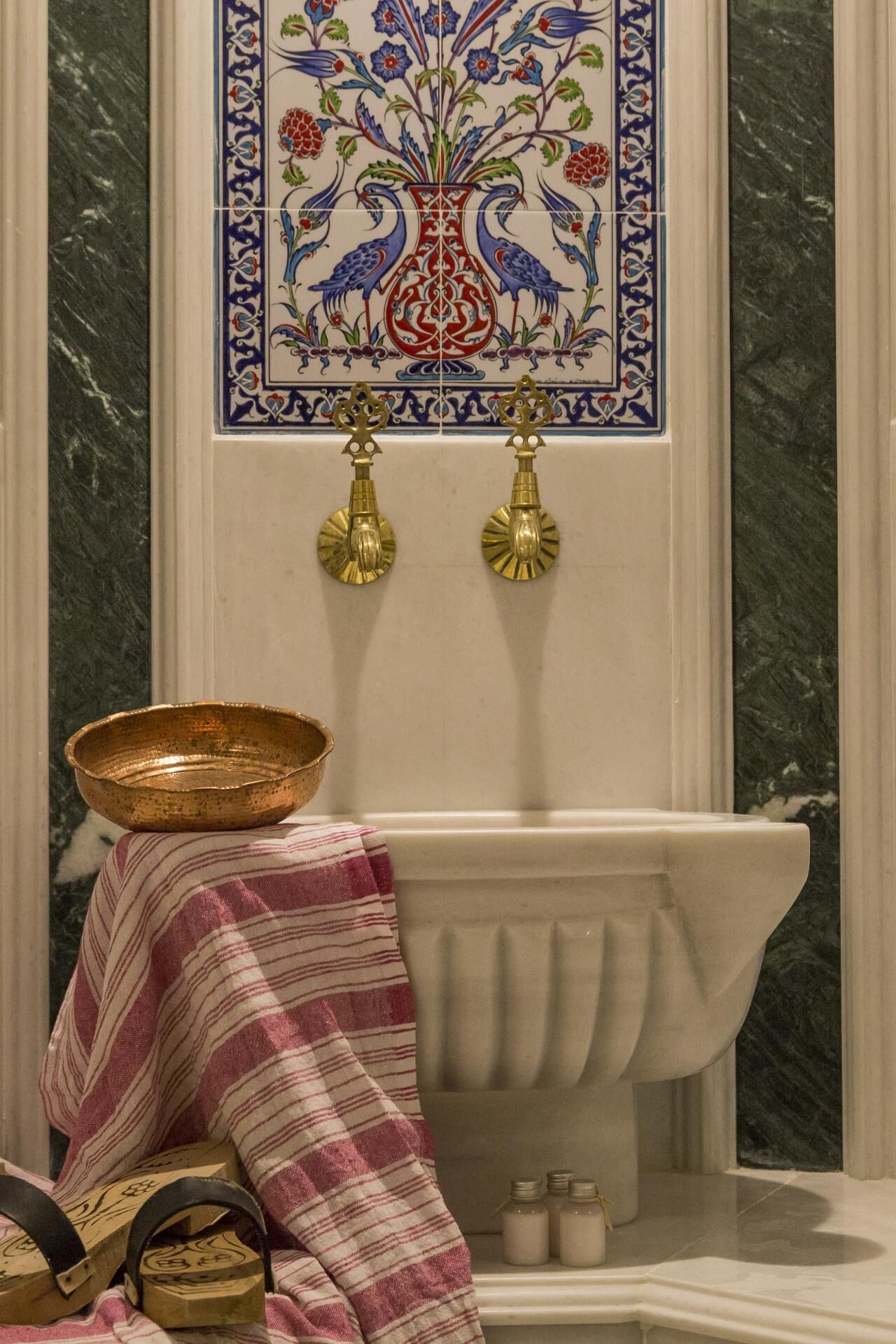 A bathroom with a marble wall, a decorative tile with peacocks, a white bathtub, and a striped cloth on a small table.