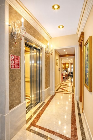 Un pasillo de hotel con suelo de mármol, un ascensor, candelabros y cuadros en las paredes.