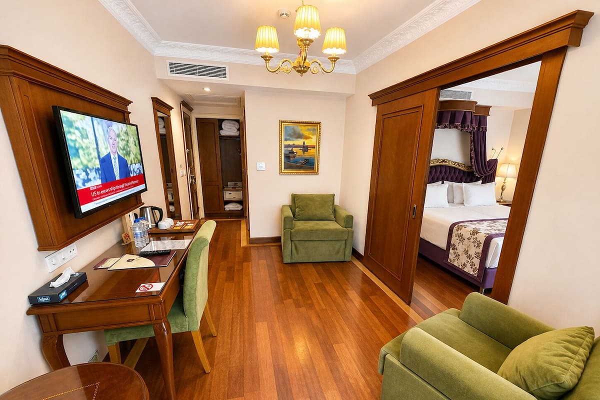 A hotel room with a wooden desk, a television, a green armchair, a bed with a purple headboard, and a painting on the wall.