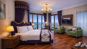 A hotel room with a bed, two green armchairs, a chandelier, a television, and a view of the sea.