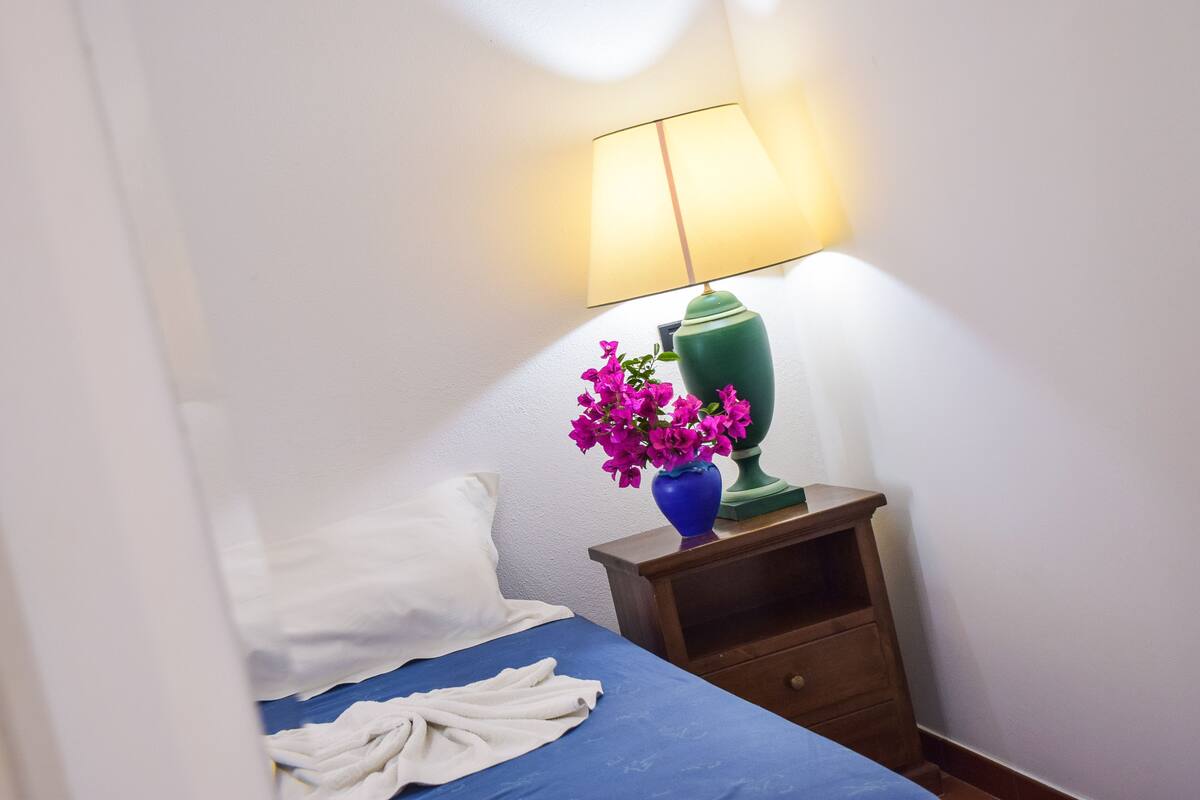 A neatly made bed with a blue bedspread and white pillows, a small wooden nightstand with a green lamp and a blue vase with pink flowers, and a white wall.