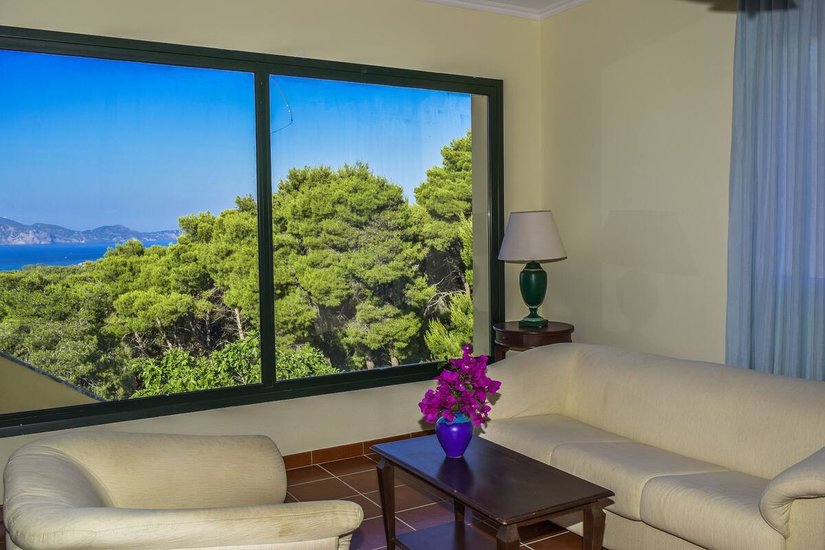 A room with a large window offering a view of a forest and the sea. There is a sofa and a small table with a vase of flowers.
