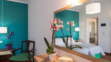 Une chambre d’hôtel aux murs de couleur turquoise, avec une chaise en bois, un bureau sur lequel se trouve un chapeau, et des orchidées.