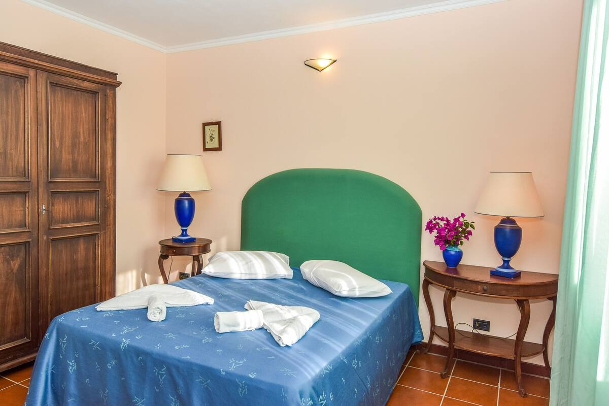 A bedroom with a bed, two bedside tables with lamps, a green headboard, and a wooden wardrobe.