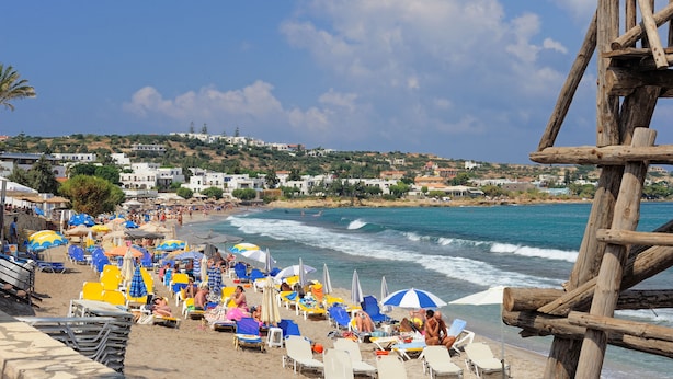 Una playa con filas de sillas de playa y sombrillas, una torre de salvavidas de madera y edificios al fondo.