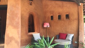 Uma área de estar coberta ao ar livre, com paredes em terracota, uma mesa pequena e um vaso com planta.