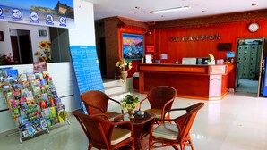 A reception area with a red desk, wicker chairs, a glass-top table, a vase with flowers, and a wall clock. - PK Mansion (Krabi)