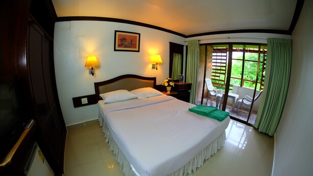 A hotel room with a large bed, two bedside lamps, a painting on the wall, and a view of greenery through the window. - PK Mansion (Krabi)