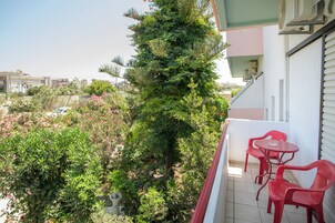 Un balcón con sillas rojas y una mesa, rodeado de vegetación exuberante y vistas de la ciudad.