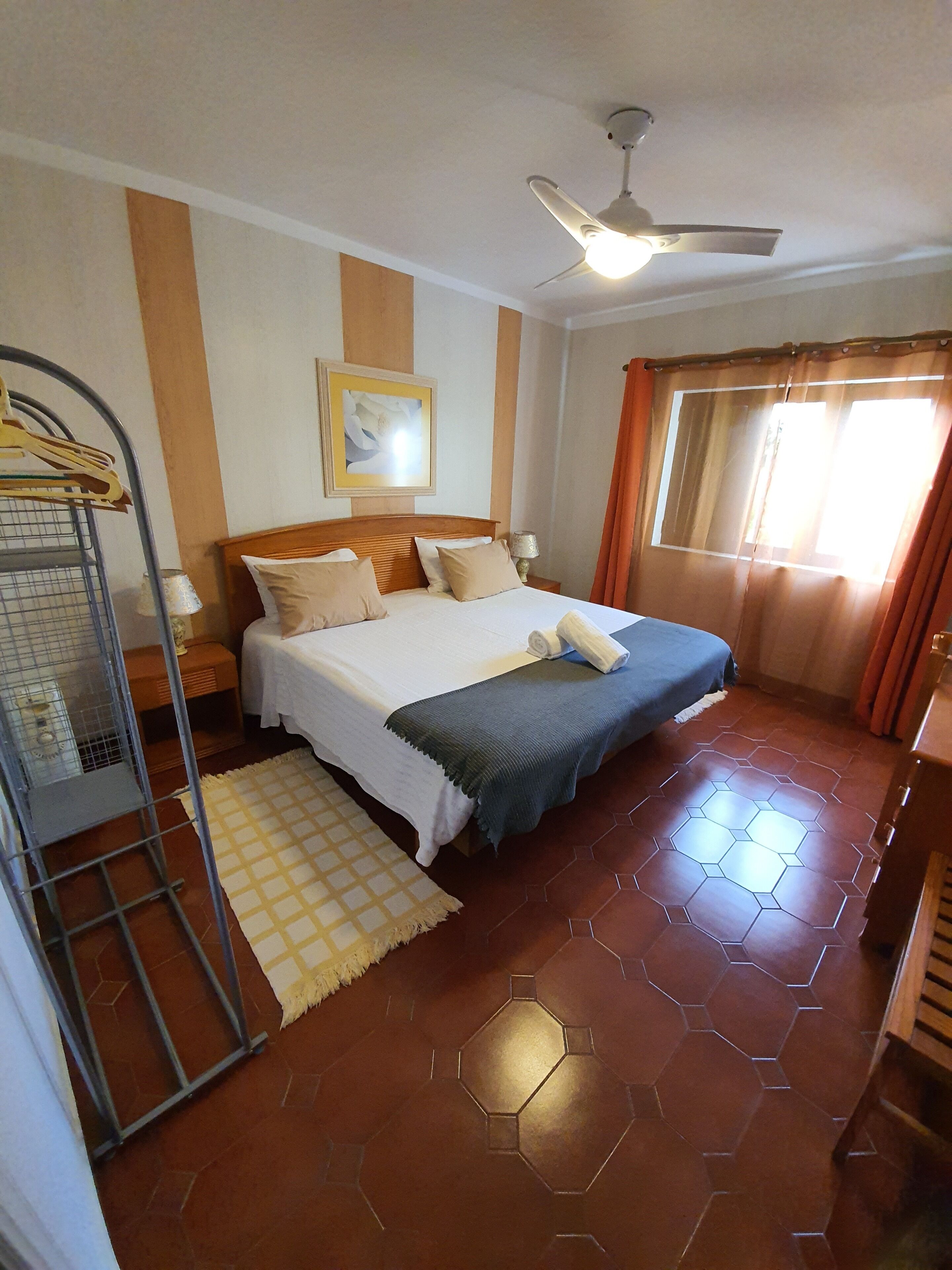 A bedroom with a bed, a ceiling fan, a window with curtains, a wall-mounted picture, and a metal rack.