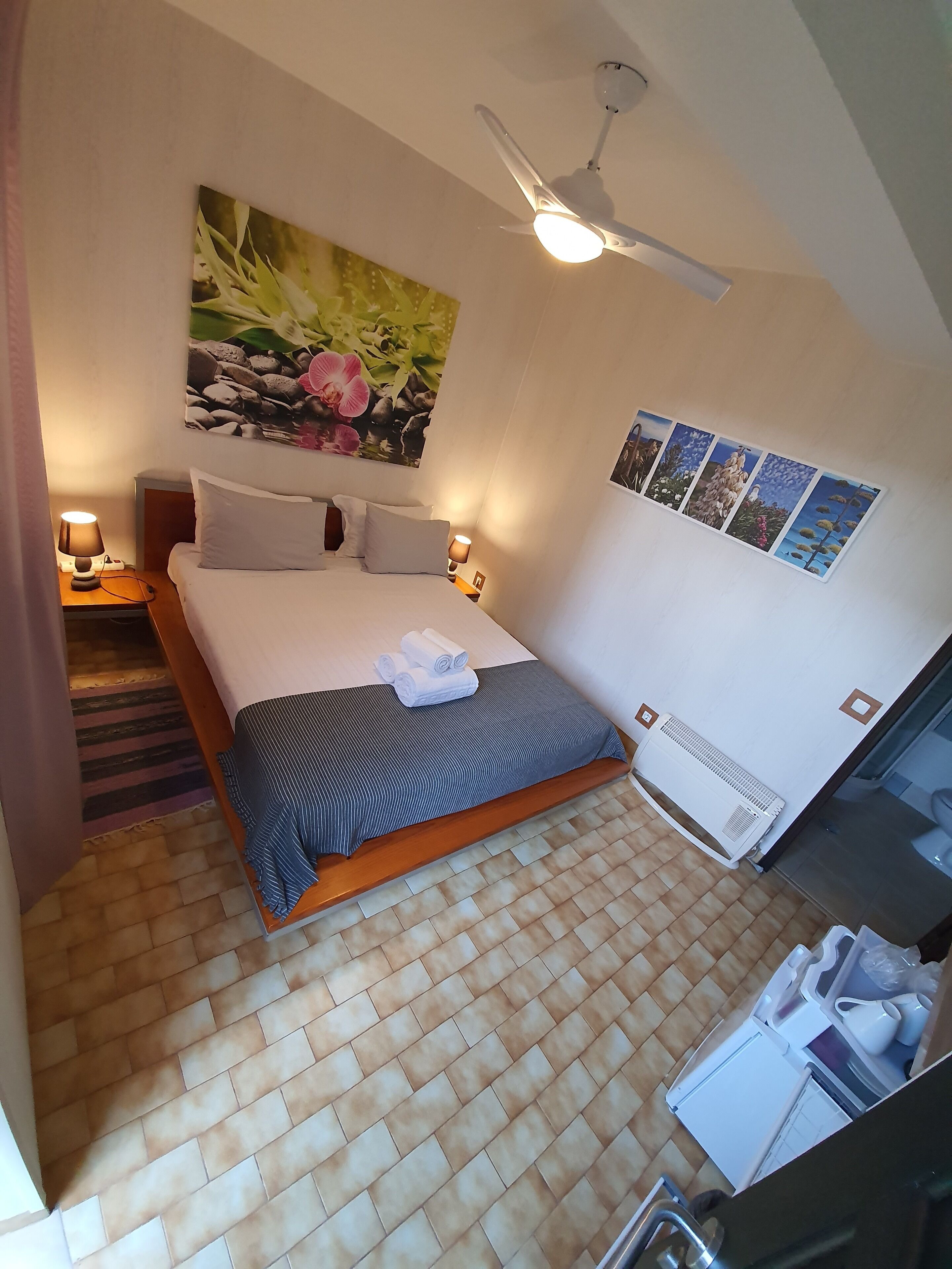 A neatly arranged bedroom with a bed, a ceiling fan, a wall-mounted air conditioner, and a series of framed pictures.