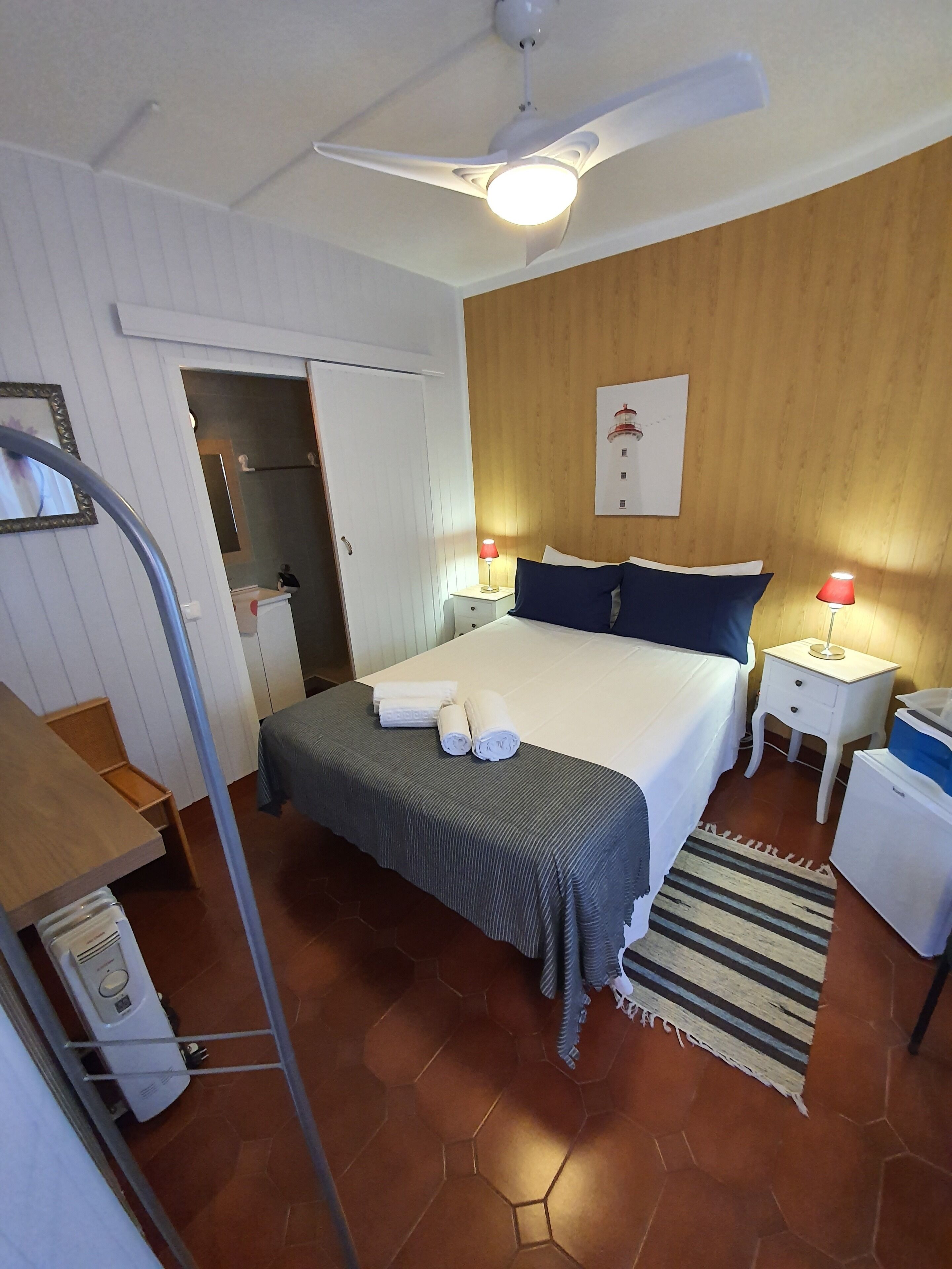 A bedroom with a bed, two bedside tables with lamps, a ceiling fan, a wooden wall panel, a tiled floor, and a door leading to a bathroom.