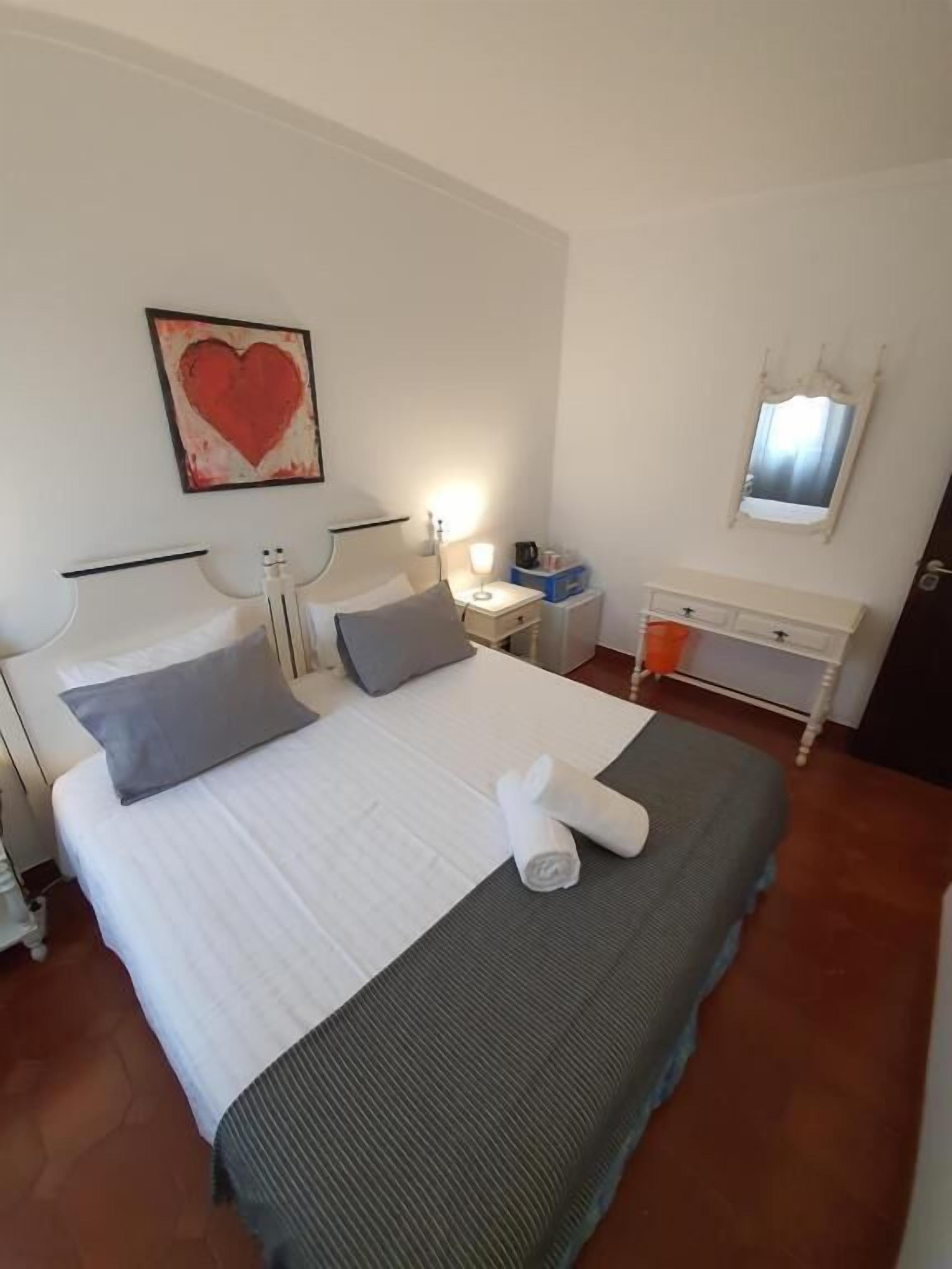 A neatly made bed with pillows, a bedside table with a lamp, a mirror, and a heart-shaped artwork on the wall.