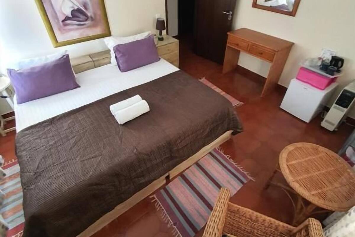 Quarto de hotel com cama, escrivaninha, cadeira, mesinha e ventilador de teto.