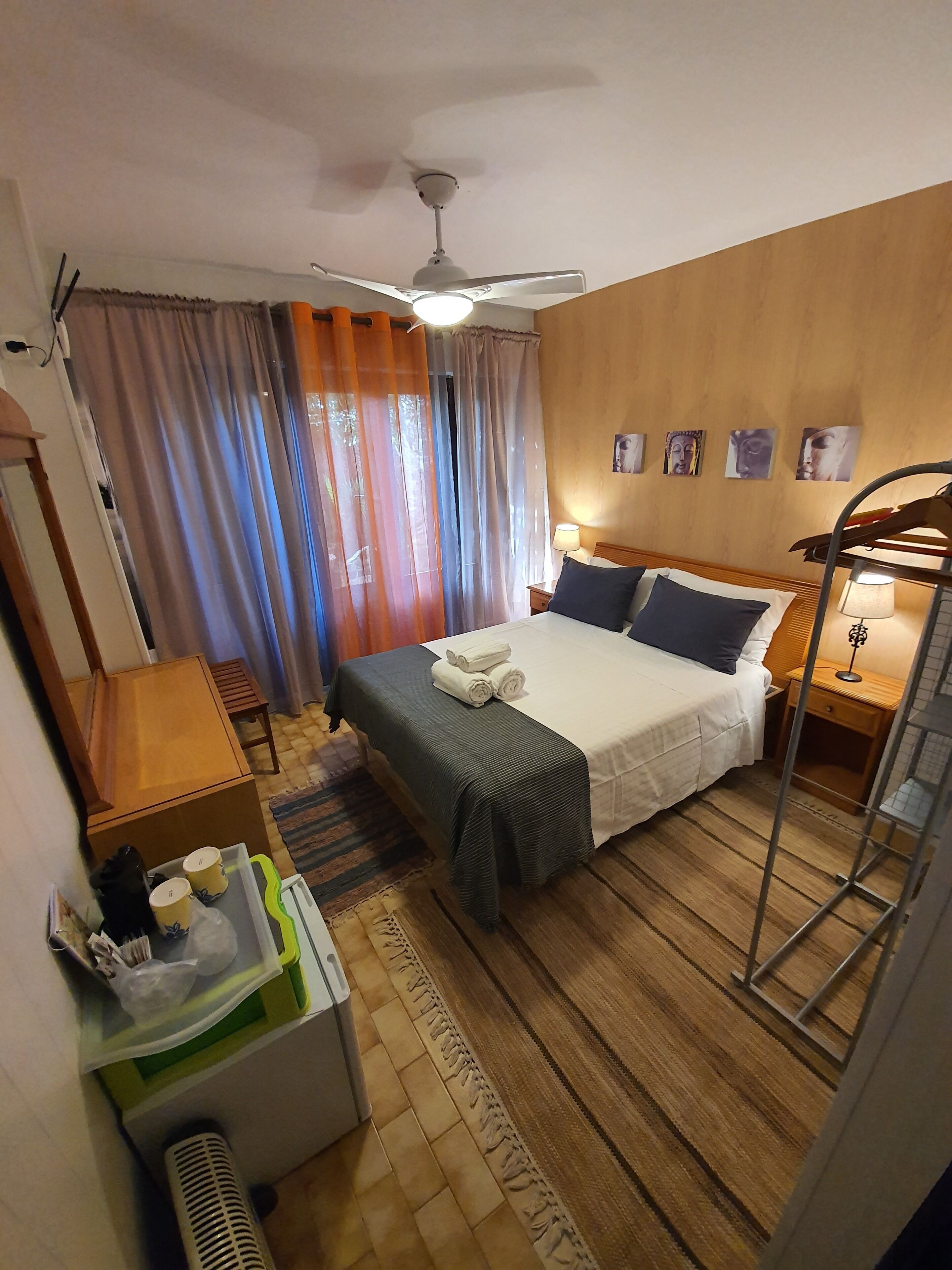 A hotel room with a bed, a ceiling fan, curtains, a wooden floor, a bedside table with a lamp, and a small shelf with toiletries.