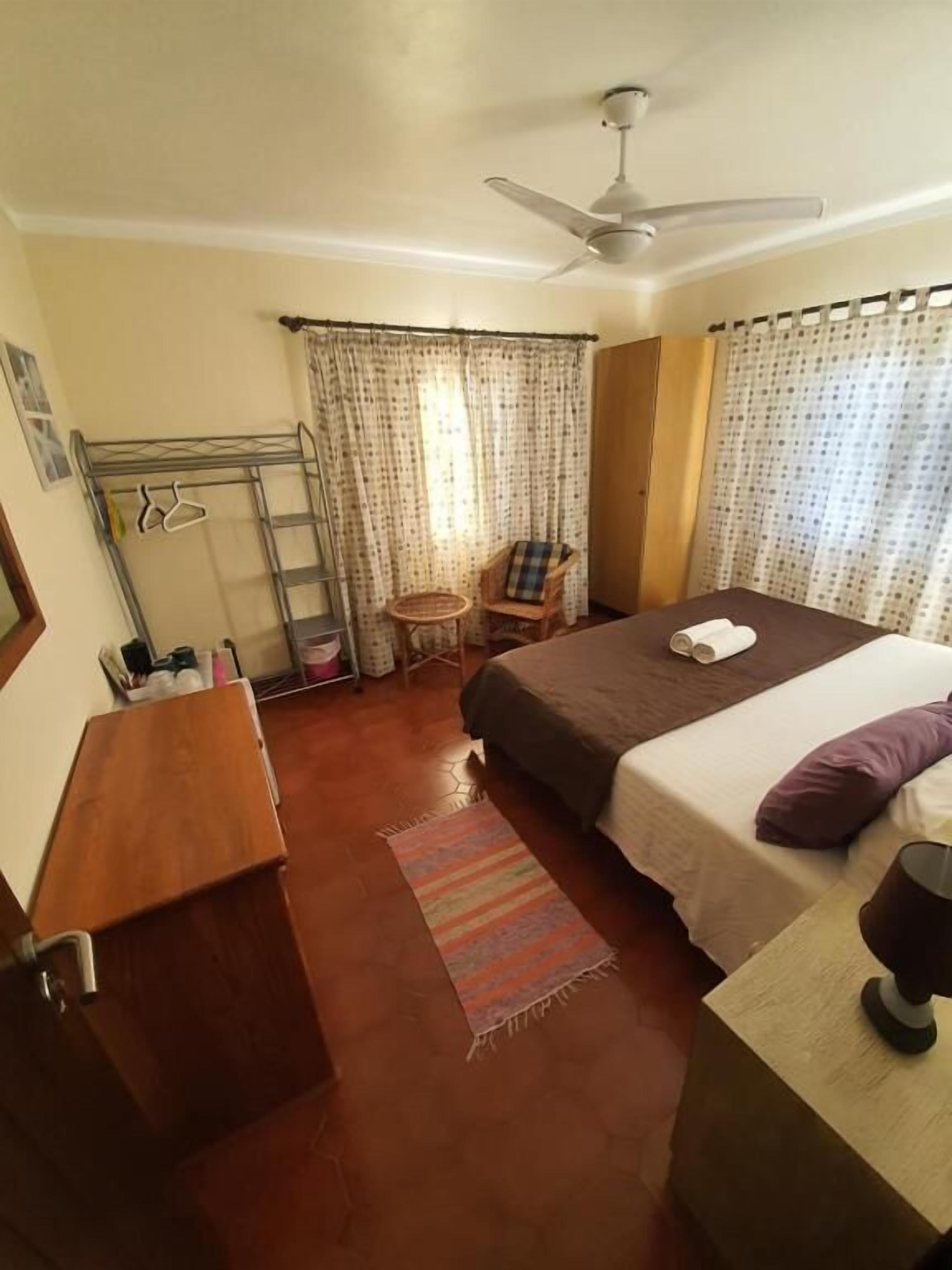 A bedroom with a bed, a chair, a small table, a ceiling fan, and curtains.