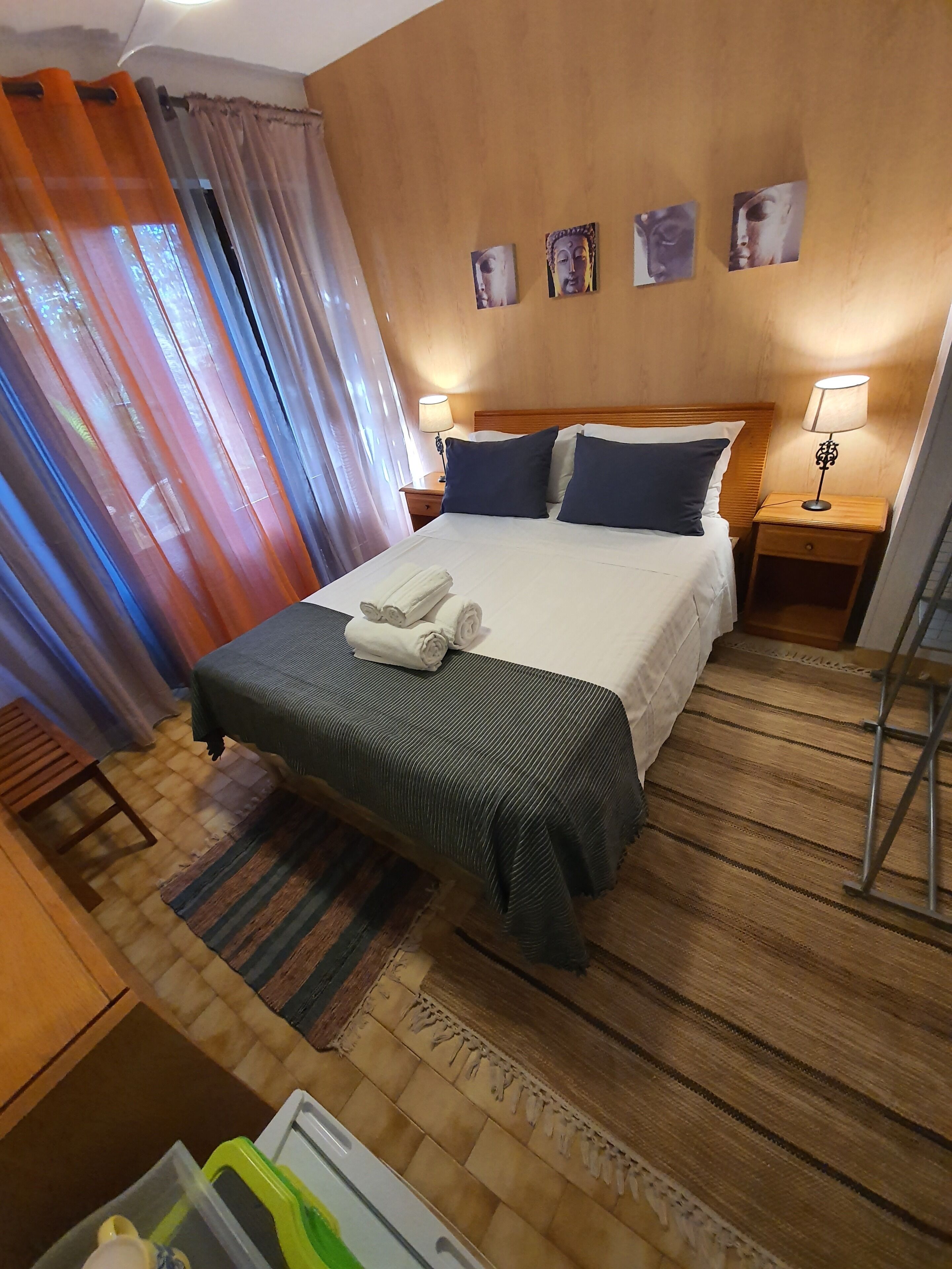 A hotel room with a bed, two bedside tables with lamps, a wooden headboard, a window with curtains, and a small table with a towel.