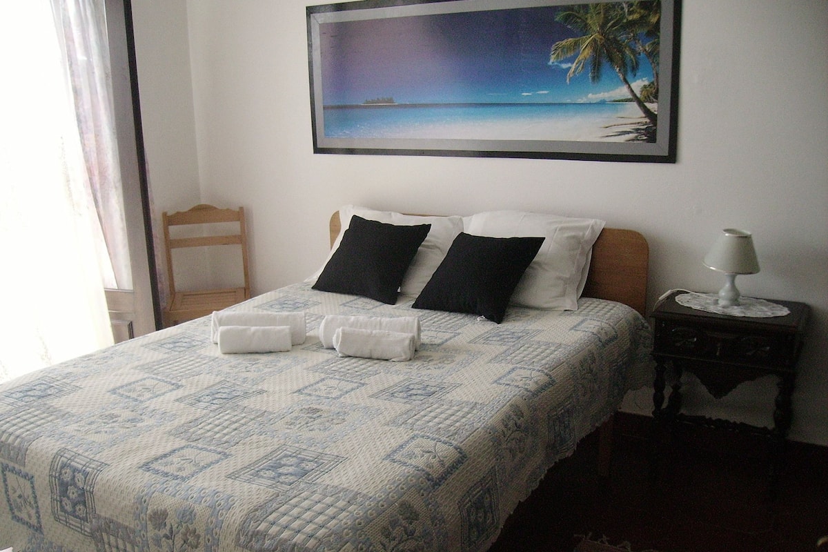 Cama bem arrumada com colcha estampada, duas almofadas pretas e uma almofada branca. Há uma moldura com uma imagem de uma praia na parede acima da cama.