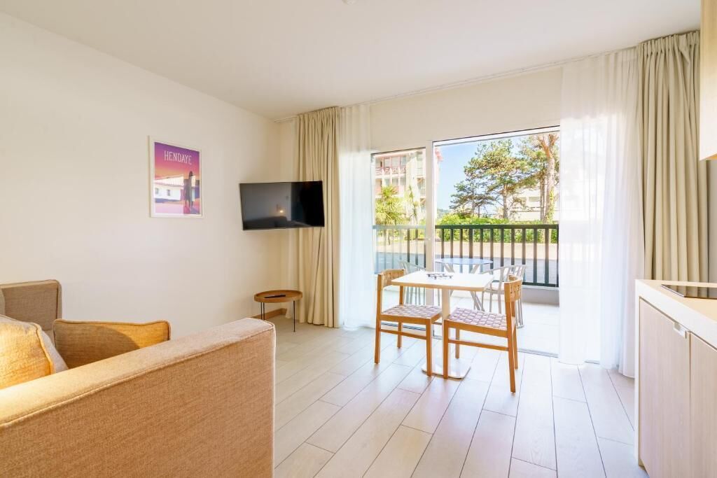 A hotel room with a sofa, a dining table with chairs, a TV mounted on the wall, and a balcony with curtains.