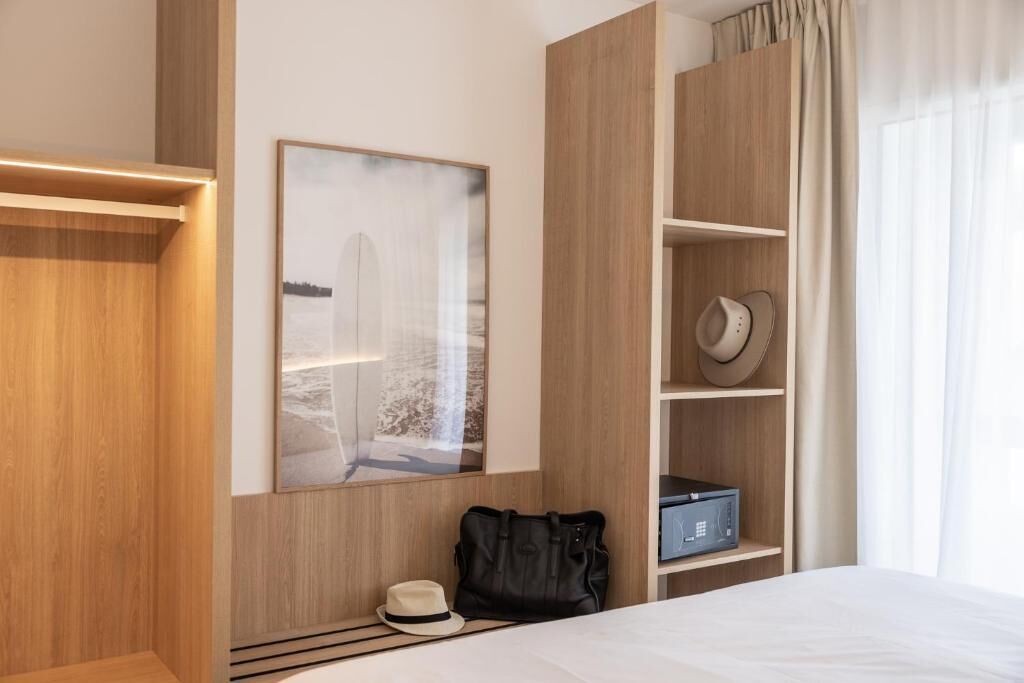 A hotel room with a bed, a bedside table, a wardrobe, a coat rack, a hat, a bag, and a framed picture on the wall.