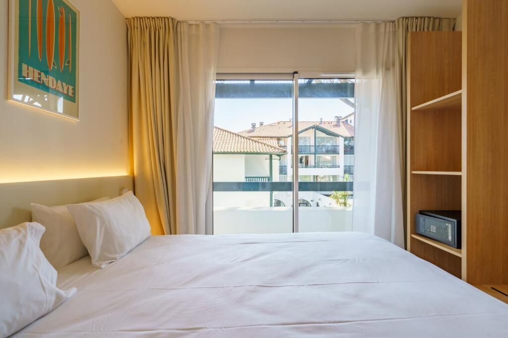 A hotel room with a bed, a view of buildings through a window, and a wooden shelf with a radio.
