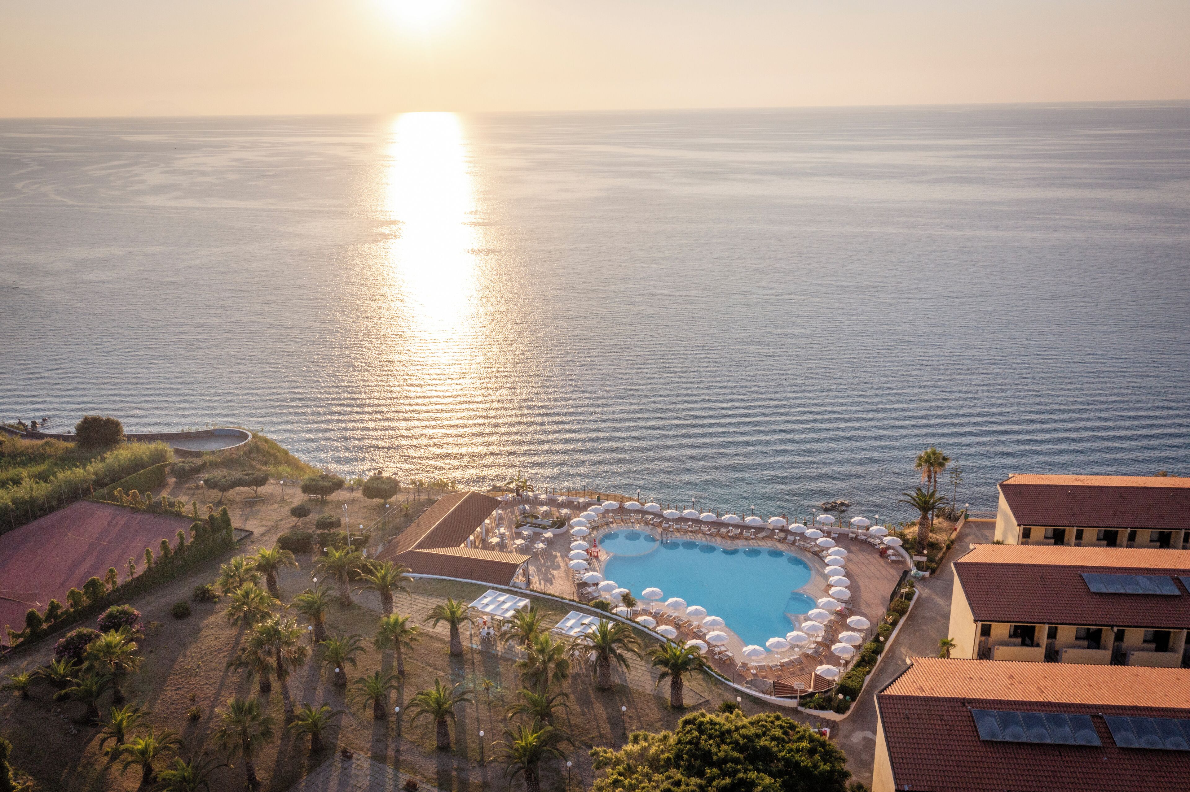 Vue aérienne d’un complexe hôtelier doté d’une piscine entourée de parasols et de palmiers, avec une vue sur la mer.