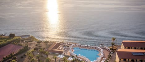 Vue aérienne d’un complexe hôtelier doté d’une piscine entourée de parasols et de palmiers, avec une vue sur la mer.