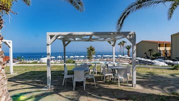 A covered outdoor seating area with white chairs and tables, overlooking a pool and beach with umbrellas.