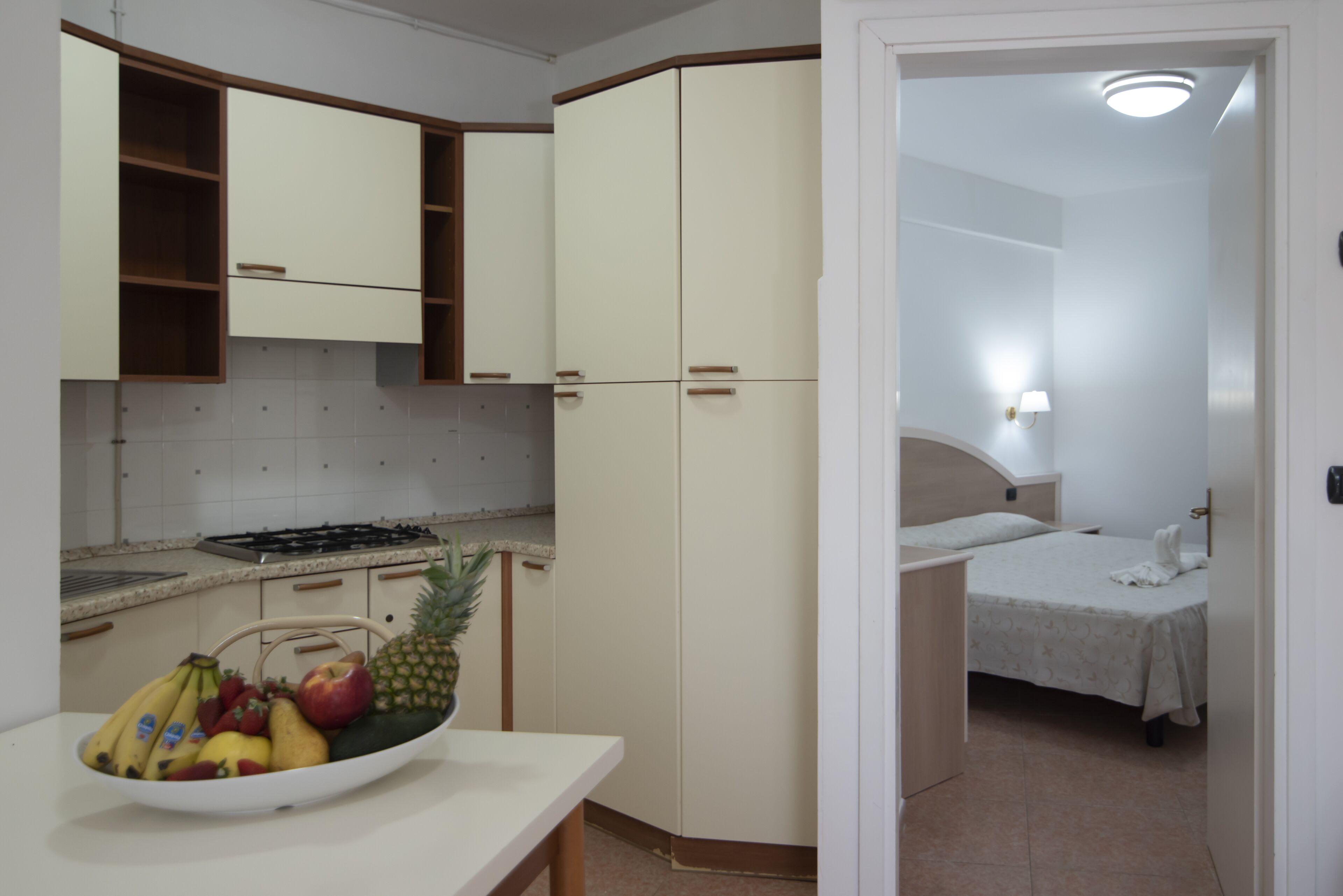 A compact kitchen with a stove, cabinets, and a fruit bowl. A bed with a nightstand and lamp is visible in the adjacent room.