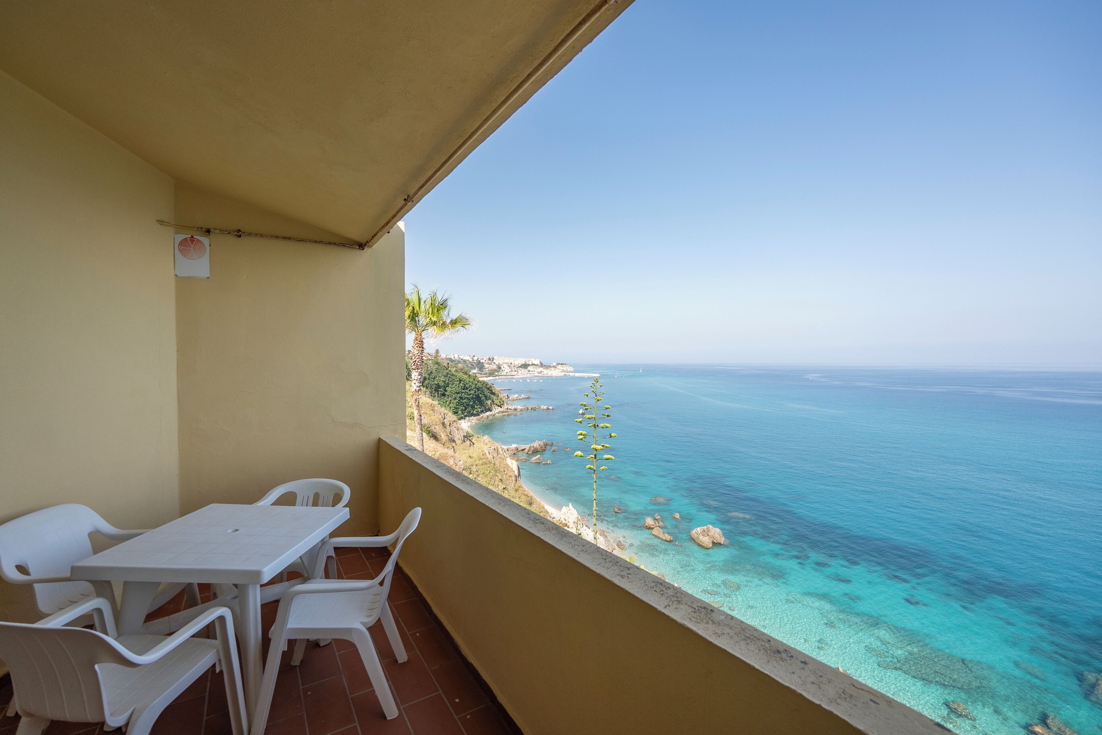 A balcony with a table and chairs, overlooking a coastal view with clear blue water and a rocky shoreline.