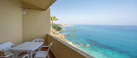 A balcony with a table and chairs, overlooking a coastal view with clear blue water and a rocky shoreline.