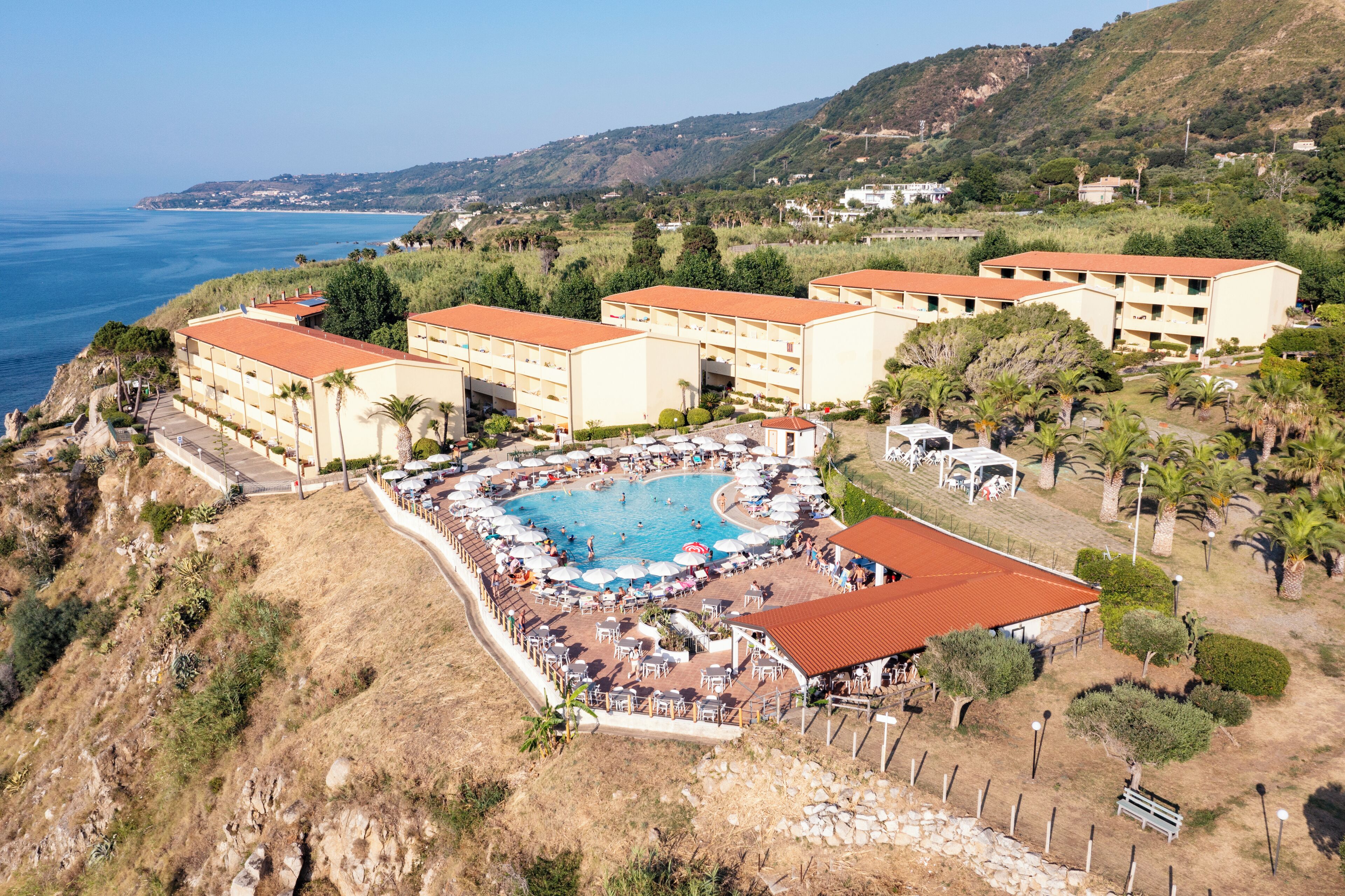 Un complexe hôtelier doté d’une piscine centrale, de plusieurs bâtiments aux toits rouges et offrant une vue sur la mer.